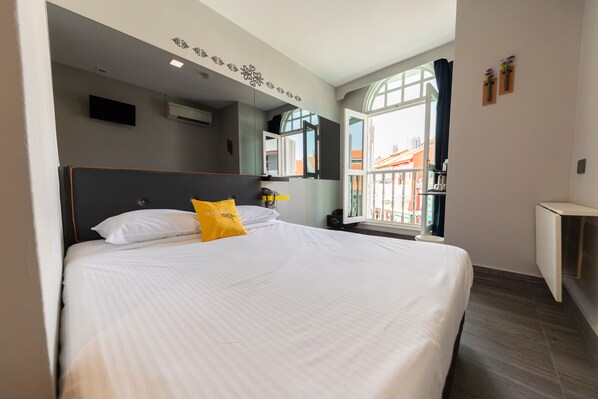 A hotel room with a large bed, a flat-screen TV, a window with a view of buildings, and a small desk with a lamp.
