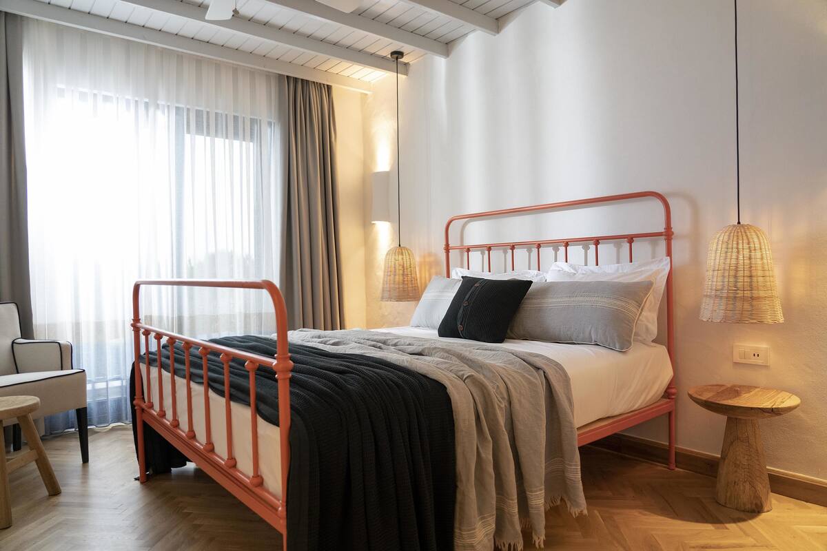 A bedroom with a metal bed frame, a bedside table, a chair, a small round table, and a window with curtains.