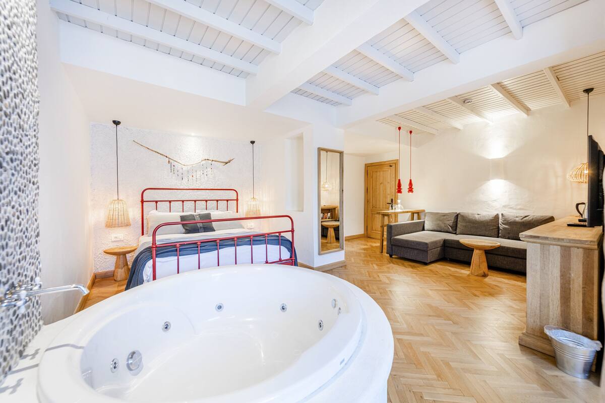 A modern bathroom with a large bathtub, a red metal bed frame, and a wooden floor.