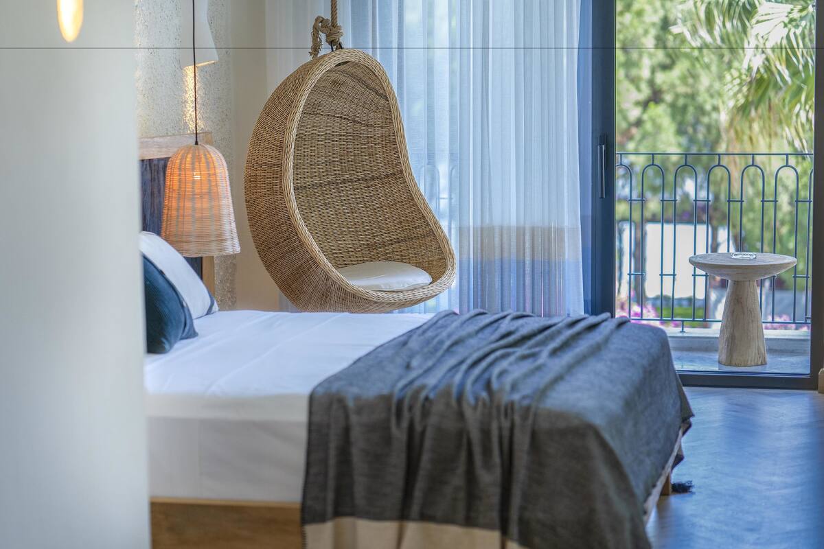 A bedroom with a bed, a hanging chair, a table, and a view of the outdoors.