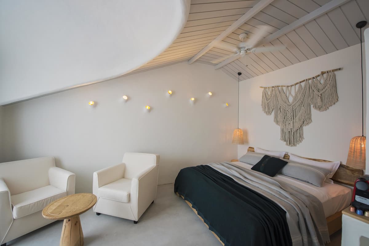 A modern bedroom with a bed, two white chairs, a small wooden table, and a ceiling fan.