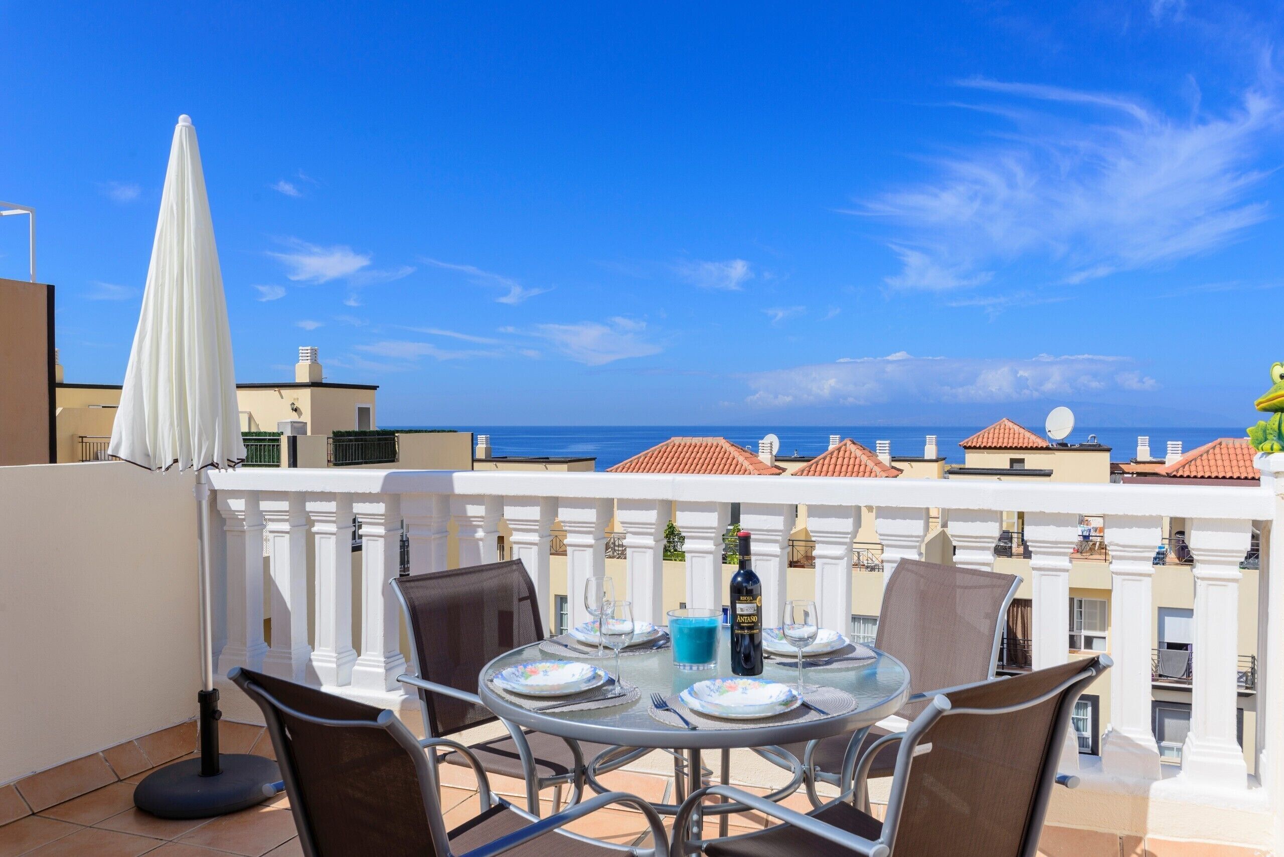 A round table with chairs on a balcony overlooking the sea, with a white umbrella and a bottle of wine on the table.