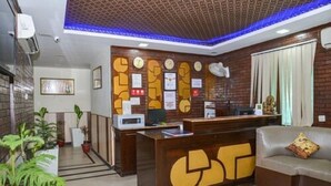 A reception desk with a clock, framed certificates, and a computer. There is a sofa with a cushion and a plant in the corner. - Airport Hotel Golden Jindal (New Delhi)