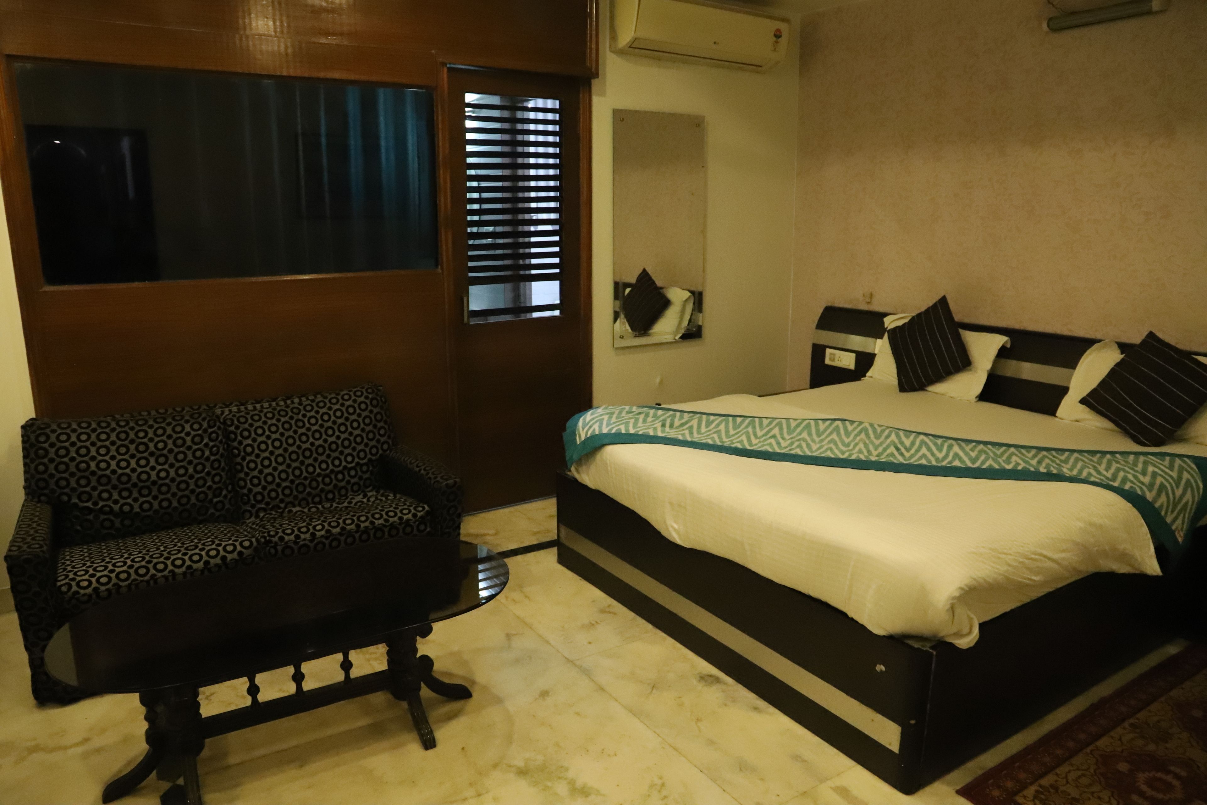 A bedroom with a bed, a sofa, a mirror, and a window with blinds.