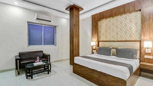 A hotel room with a large bed, a wooden headboard, a small table with a bottle, a couch, and an air conditioning unit on the wall. - Airport Hotel Golden Jindal (New Delhi)