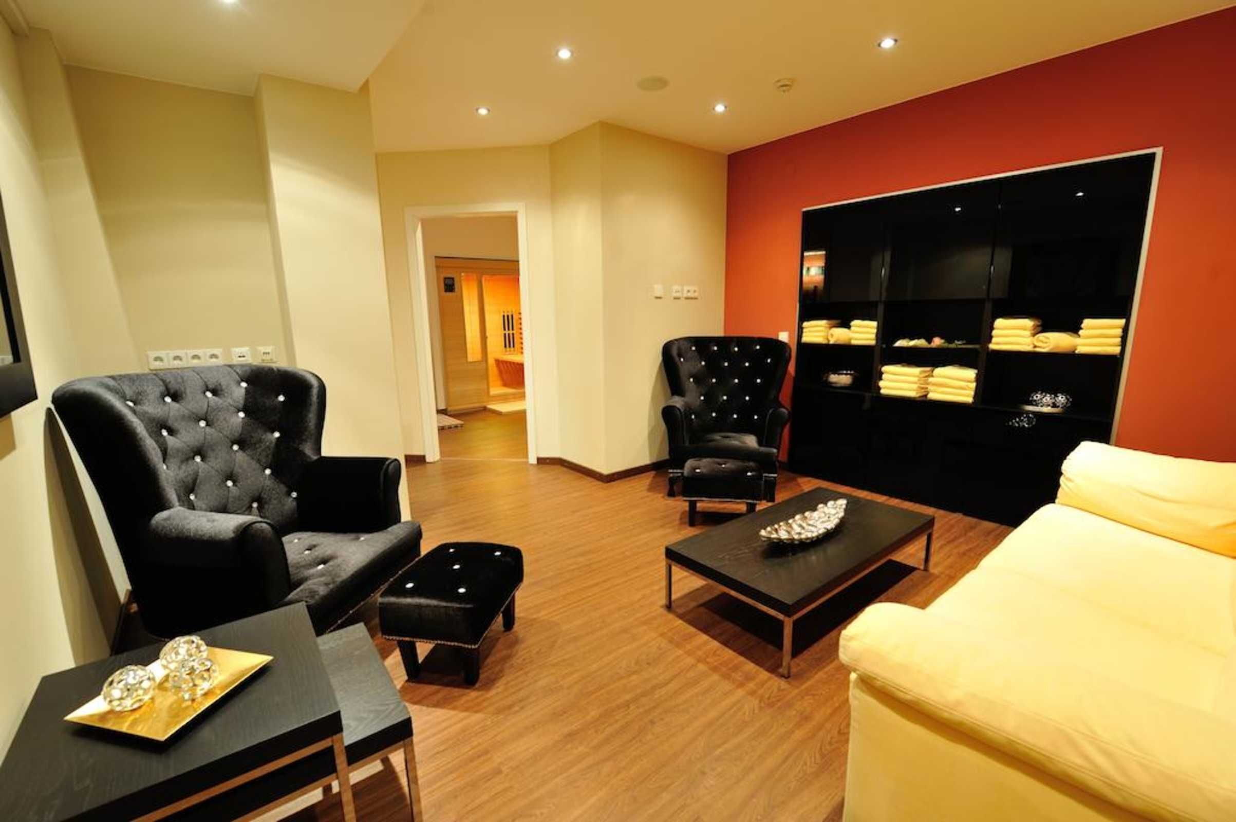 Ein modernes Wohnzimmer mit einem schwarzen, gepolsterten Sessel, einem gelben Sofa und einem weiteren schwarzen Sessel. Ein Holztisch mit einem dekorativen Gegenstand und einem schwarzen Hocker steht im Raum. An der Wand befindet sich ein schwarzes Regal mit gefalteten Handtüchern.
