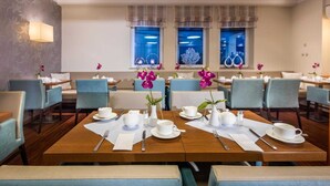 Ein Hotel-Speisesaal mit mehreren Tischen und Stühlen, jeder Tisch ist mit weißem Geschirr und violetten Orchideen dekoriert.