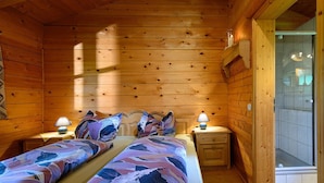 Een houten slaapkamer met twee bedden, bedlampjes en een zichtbare badkamer door een glazen deur.
