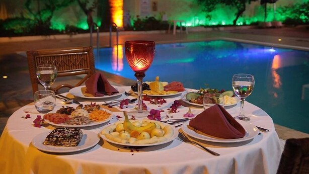 Mesa de comedor preparada para una comida junto a una piscina, con mantel, platos de comida, vasos y una vela.