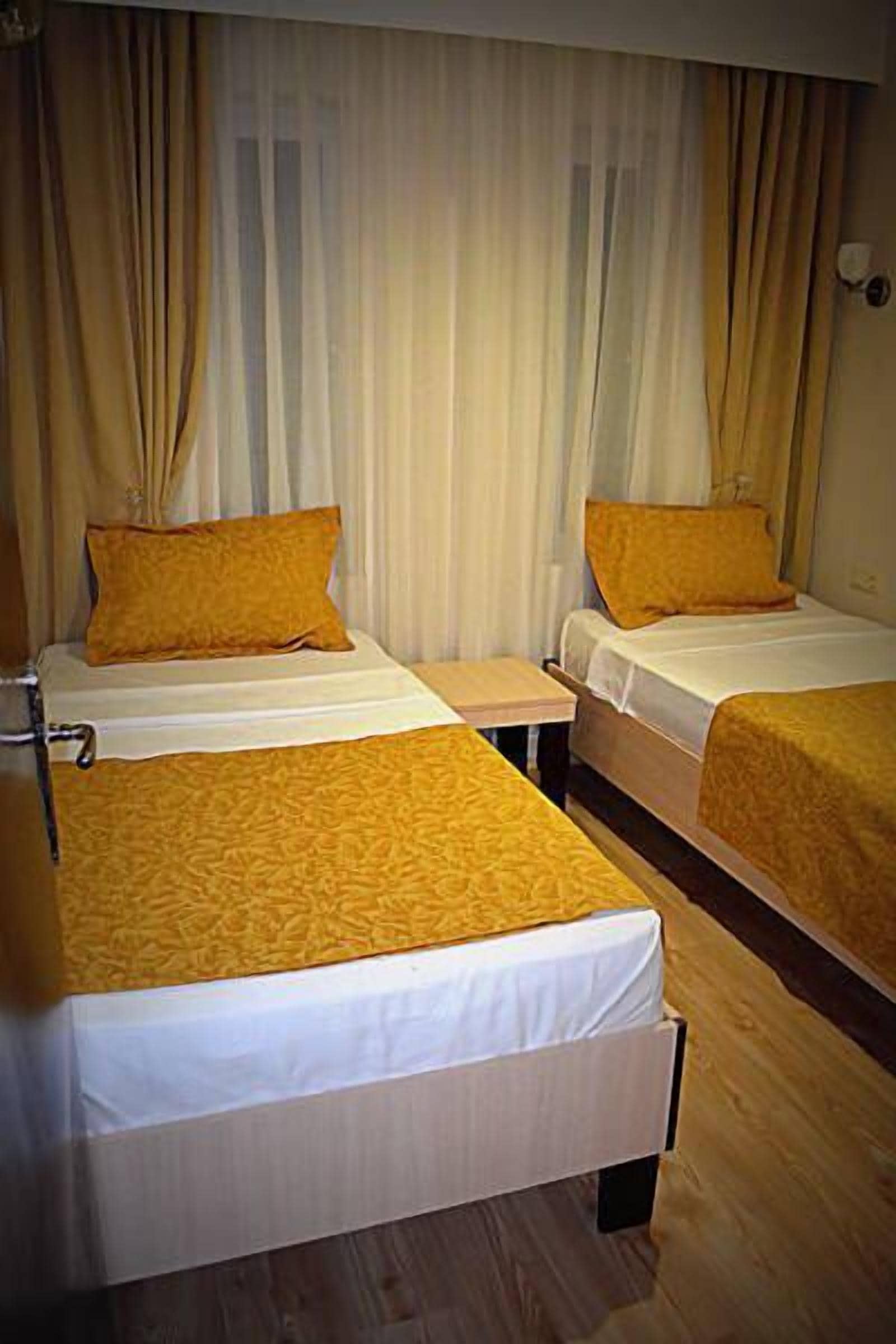 A hotel room with two beds, each with a white mattress and a yellow blanket. The room has wooden flooring and beige curtains.