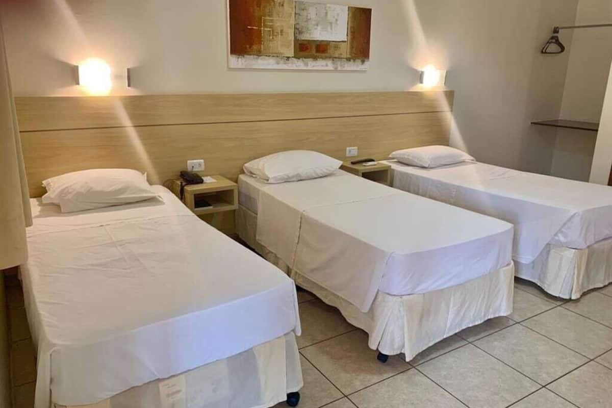 Une chambre d’hôtel avec trois lits simples, une tête de lit en bois, une œuvre d’art accrochée au mur et une table de chevet avec une lampe.