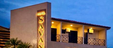 A hotel named 'Savana' with a modern design, featuring a prominent vertical sign, balconies, and well-lit surroundings.