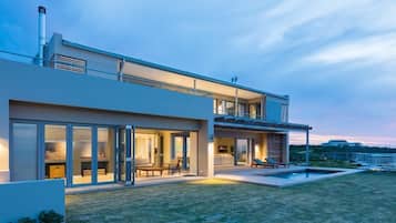 A modern two-story house with a large glass facade, outdoor seating area, and a swimming pool.