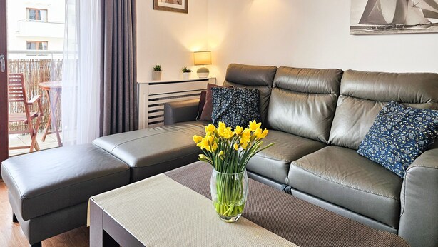 A modern living room with a grey leather sofa, a small side table, and a vase with yellow flowers. - 2-room WO07 - Platan (WO07) (Swinoujscie)