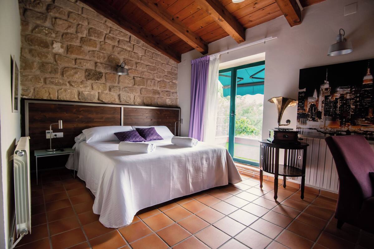 Un dormitorio con pared de piedra, vigas de madera en el techo, una cama con ropa blanca, una mesita de noche, una ventana con cortinas moradas y vistas a la ciudad.