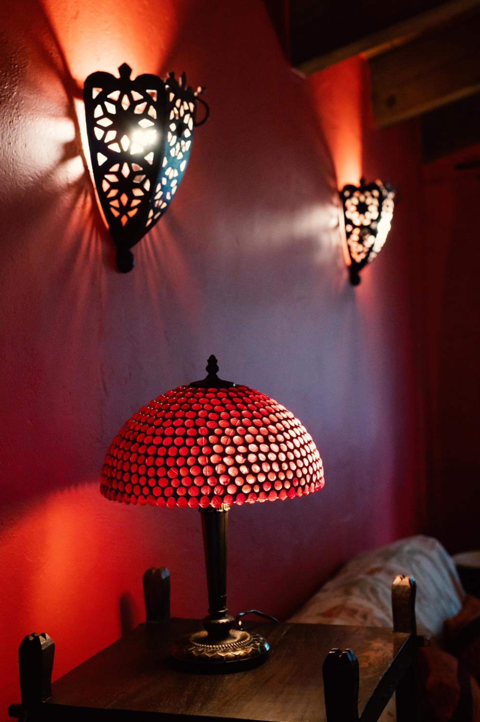 Una lámpara de mesa con una pantalla roja a lunares sobre una mesa de madera, con dos lámparas fijadas a la pared al fondo.