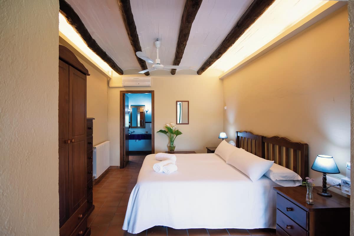 Habitación de hotel con una cama grande, una mesita de noche con lámpara, un armario de madera, un espejo y un ventilador de techo.
