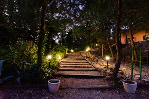 A stone pathway leading through a garden at dusk, flanked by trees and illuminated by street lamps. - Hospedium Hotel la Parada del Compte (Torre del Compte)