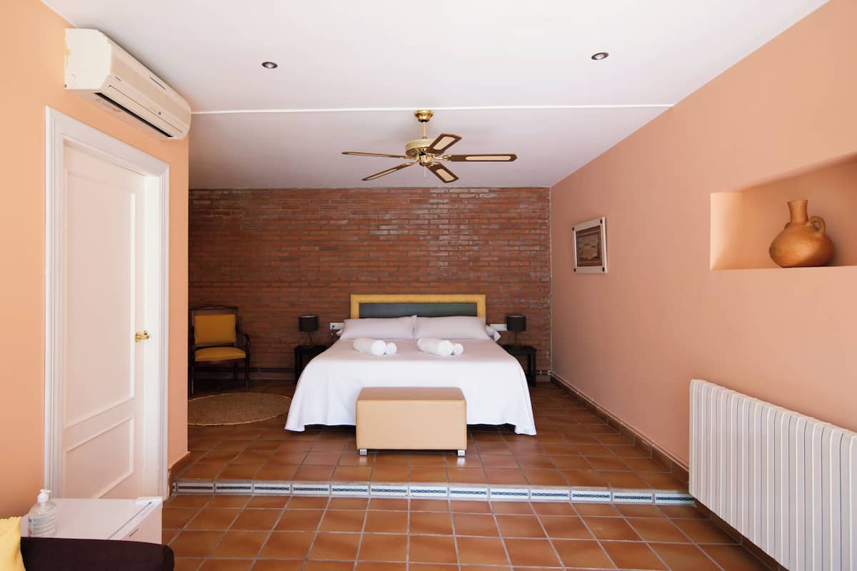 Un dormitorio con pared de ladrillo, una cama con ropa blanca, un ventilador de techo, una mesita de noche con lámpara y un radiador.