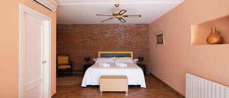 A bedroom with a brick wall, a bed with white linens, a ceiling fan, a bedside table with a lamp, and a radiator.