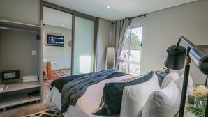 A modern hotel room with a bed, a desk, a TV, and a window with curtains. - Suite 9 The Village (Windhoek)