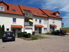 A multi-story residential building with a red-tiled roof, a balcony with a yellow umbrella, and two cars parked in the driveway.