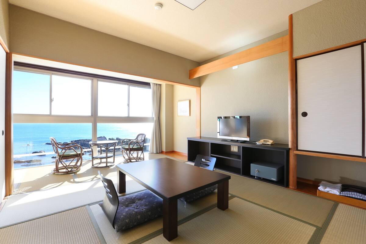 A room with a large window overlooking the sea, a wooden table, chairs, a television, and a wardrobe.
