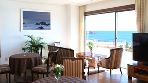 Une chambre d’hôtel dotée d’une grande fenêtre donnant sur la mer, d’un téléviseur posé sur un meuble en bois et d’un coin repas avec une table et des chaises.