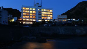 Un hôtel à plusieurs étages avec des fenêtres illuminées, une rivière au premier plan et un paysage montagneux en arrière-plan.
