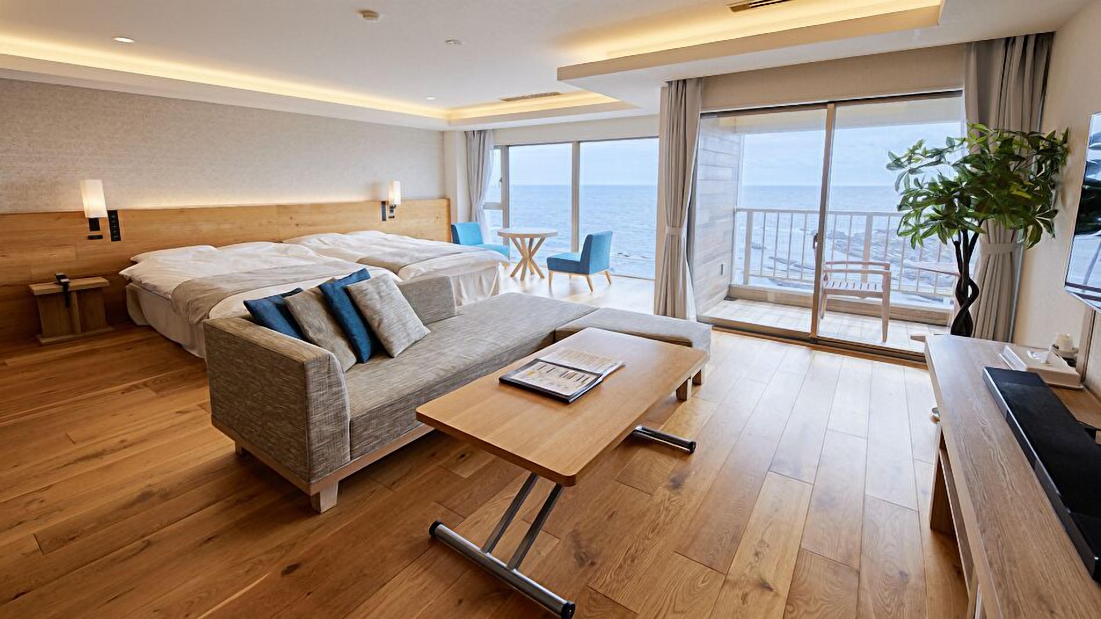 A modern hotel room with a wooden floor, a sofa, a bed, a small table, a chair, and a balcony with a view of the sea.