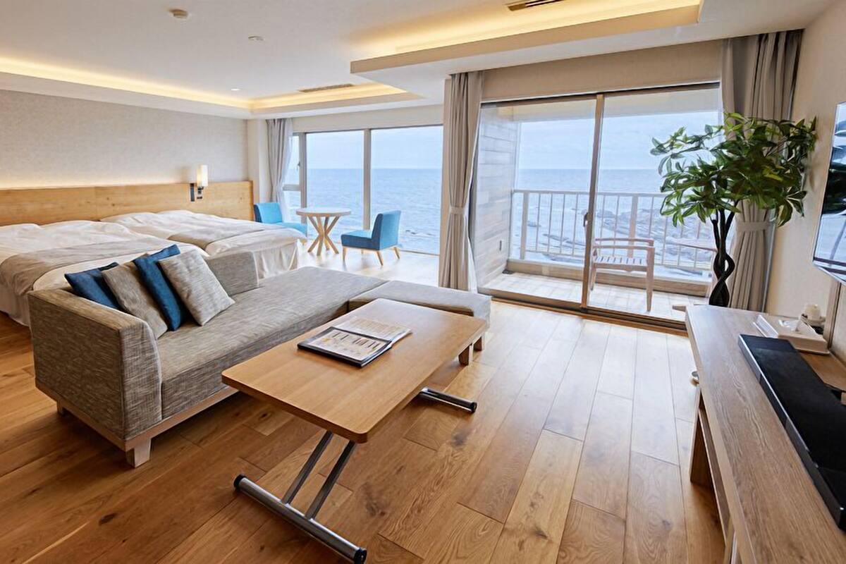 A modern hotel room with a wooden floor, a sofa, a bed, a small table, a chair, and a balcony with a view of the sea.