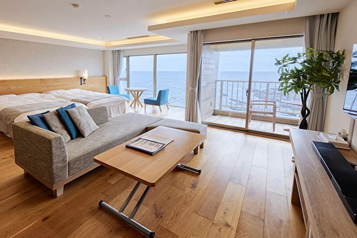Habitación de hotel moderna con suelo de madera, sofá, cama, mesita, silla y balcón con vista al mar.