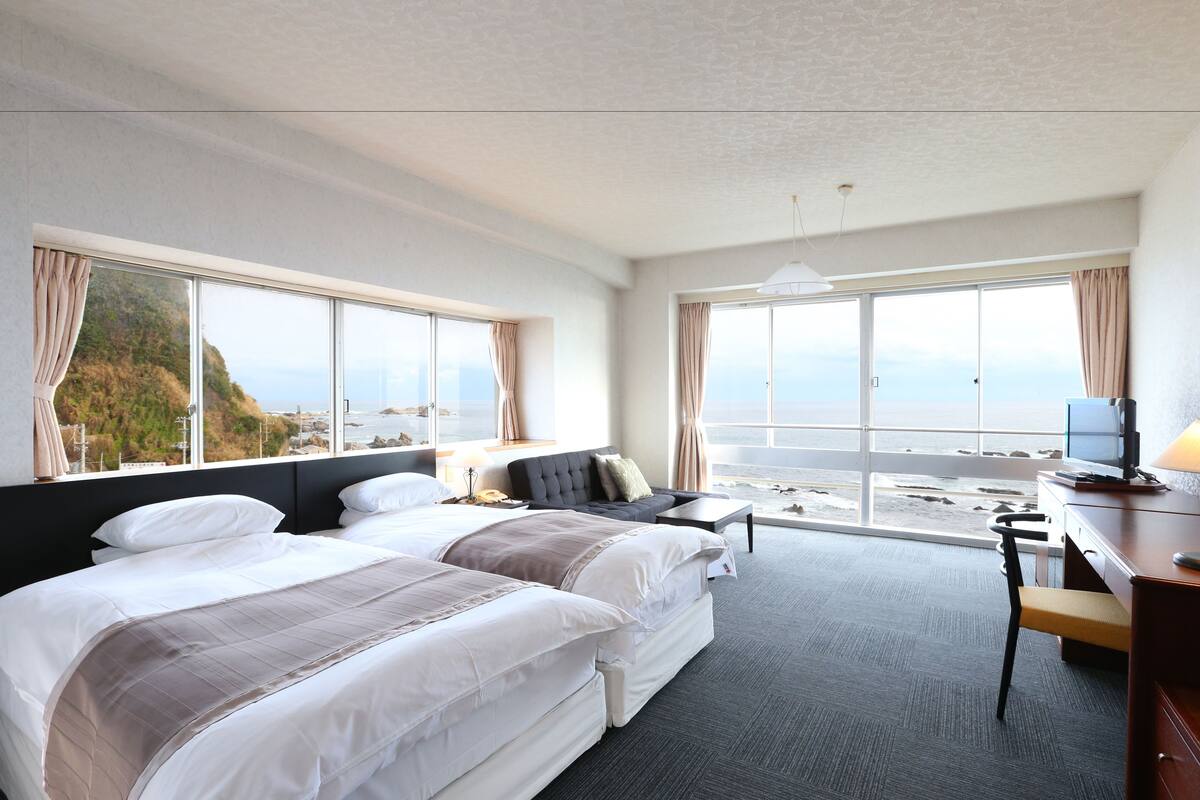 A hotel room with two beds, a desk, a chair, and large windows offering a view of the sea.