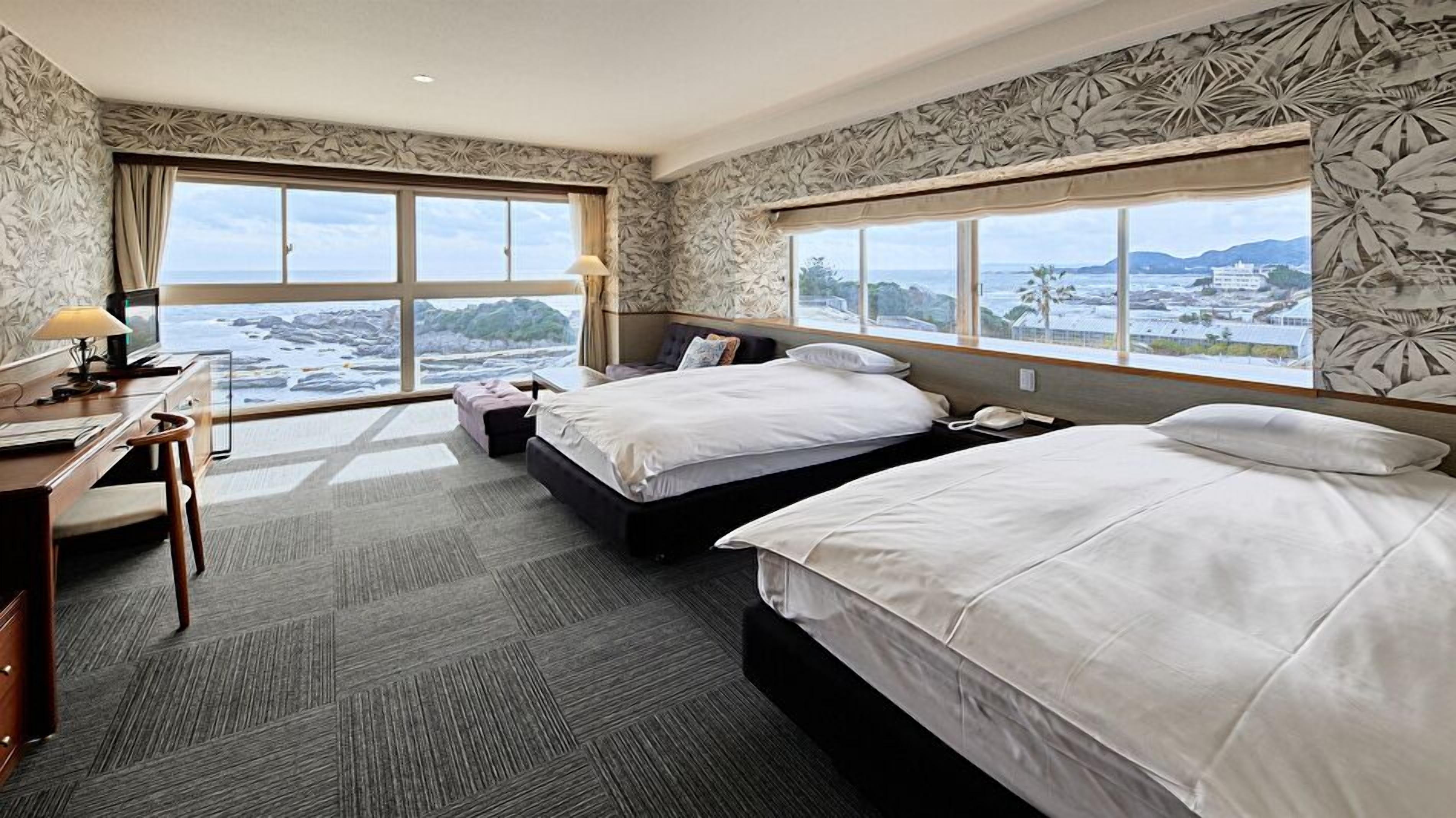 A hotel room with two beds, a desk, and large windows offering a view of the sea and mountains.