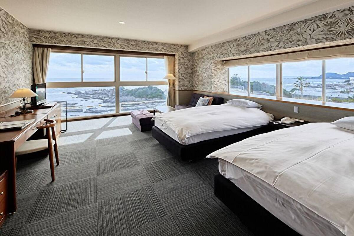 A hotel room with two beds, a desk, and large windows offering a view of the sea and mountains.