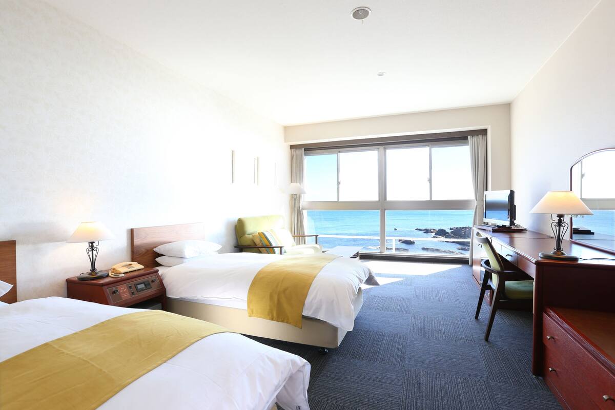 A hotel room with two beds, a desk, a chair, and a large window offering a view of the ocean.
