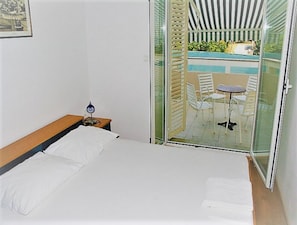 A bedroom with a bed, a nightstand, a lamp, and a sliding glass door leading to a balcony with a pool and outdoor seating. - Apartments Vrtlici (Novalja)