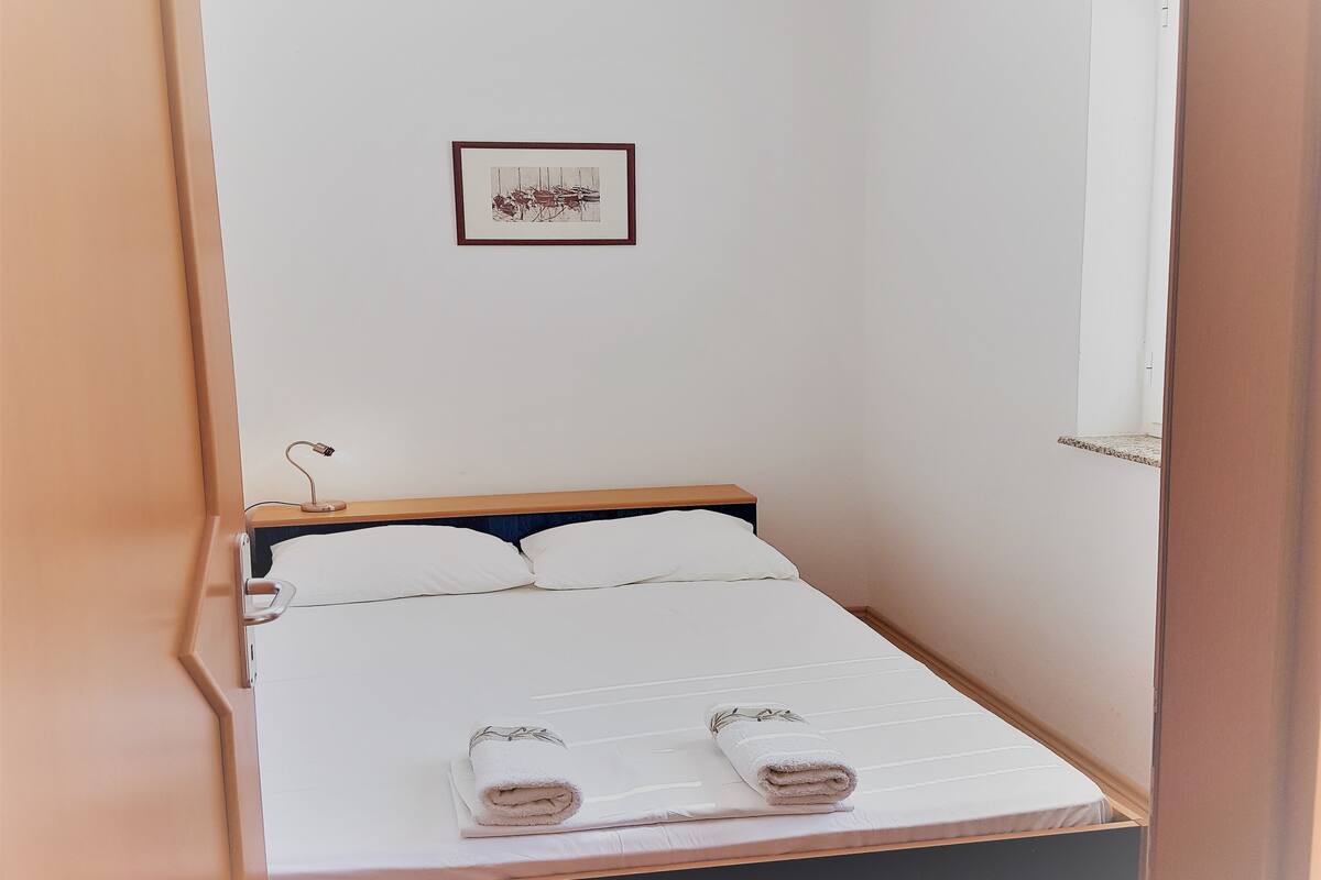 A small, neatly made bed with white linens and two rolled towels, a bedside table with a lamp, and a framed picture on the wall.