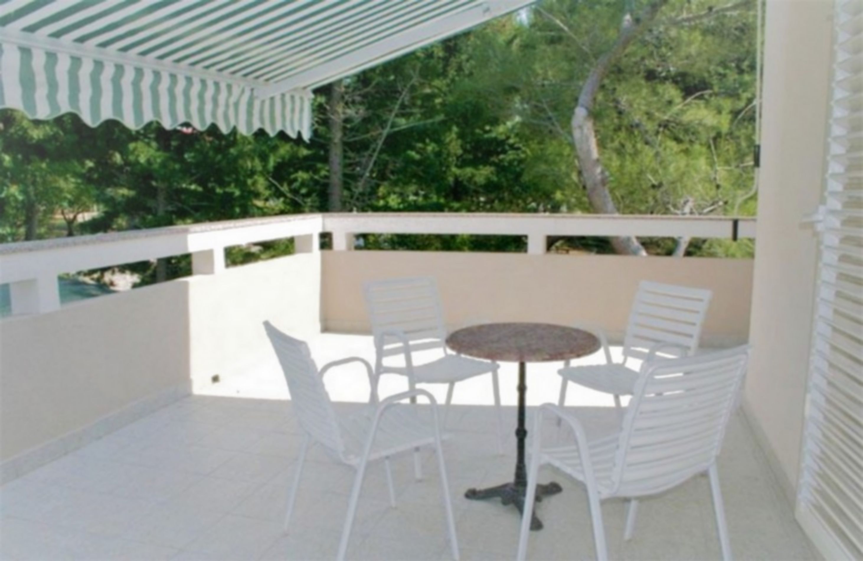 A covered outdoor area with a white metal table and chairs, surrounded by a white railing, and trees in the background.