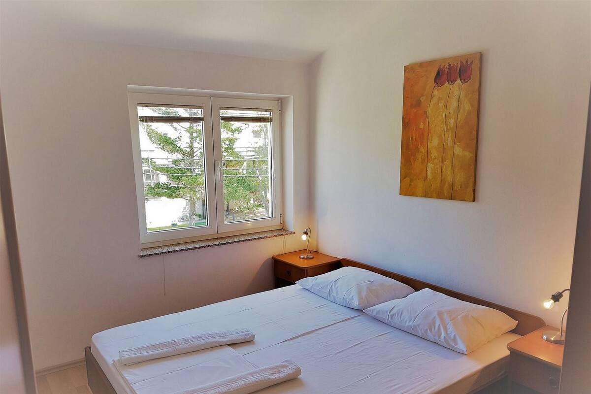 A bedroom with a single bed, a window with blinds, a bedside table with a lamp, and a painting on the wall.