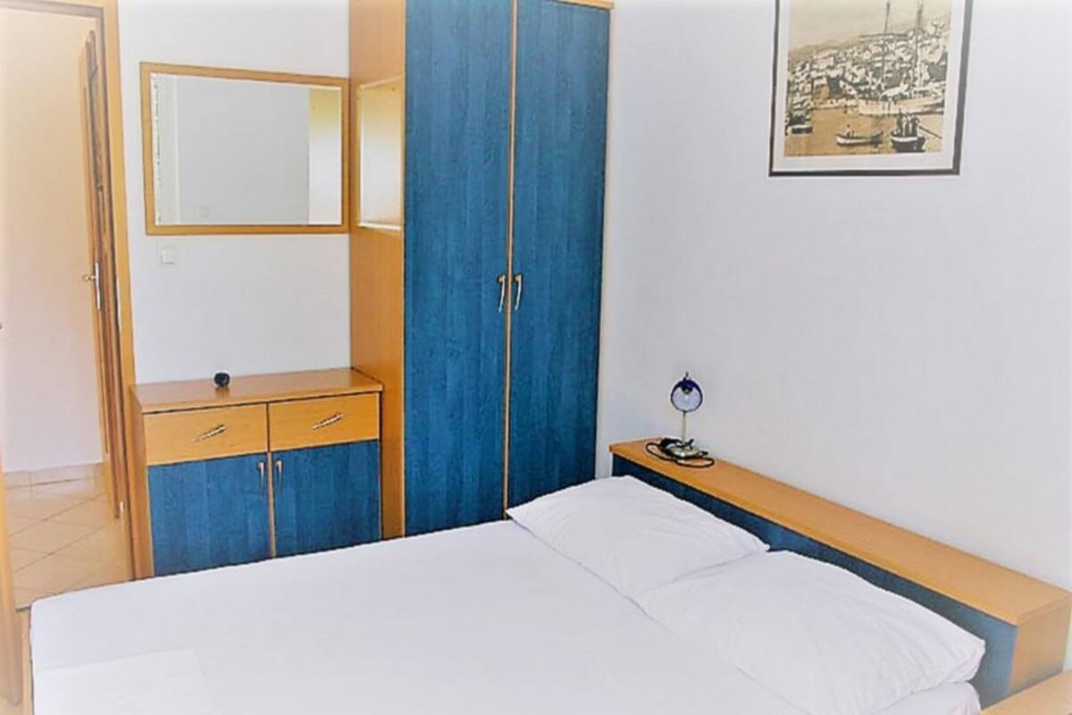 A bedroom with a bed, a wooden dresser, a blue wardrobe, and a bedside table with a lamp.