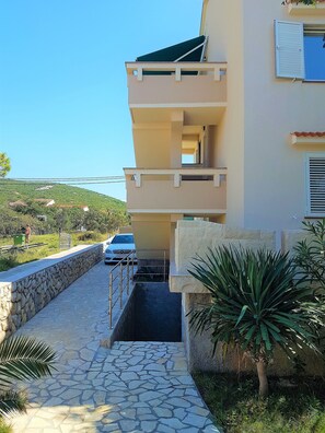 A multi-story building with a stone pathway leading to the entrance, surrounded by greenery and a hillside in the background. - Apartments Vrtlici (Novalja)