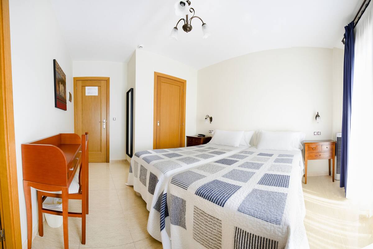 Quarto de hotel com cama, secretária, cadeira, guarda-roupa, mesa de cabeceira e luz no teto.