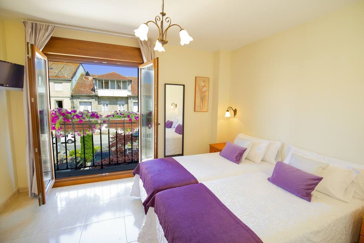 Quarto de hotel com duas camas, varanda com vista para prédios e flores, e porta de vidro deslizante.
