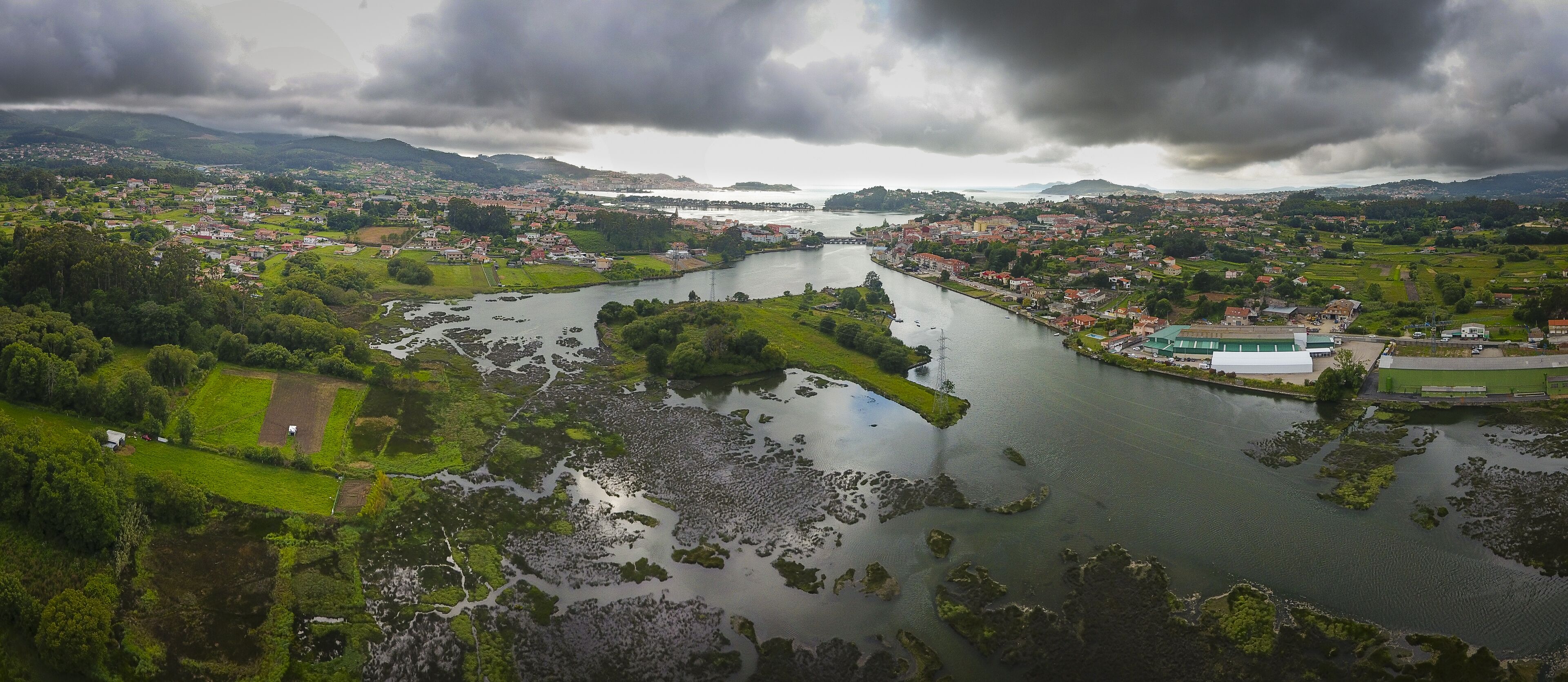 An aerial view of a river flowing through a town with green fields, a bridge, and a large building with a green roof.
