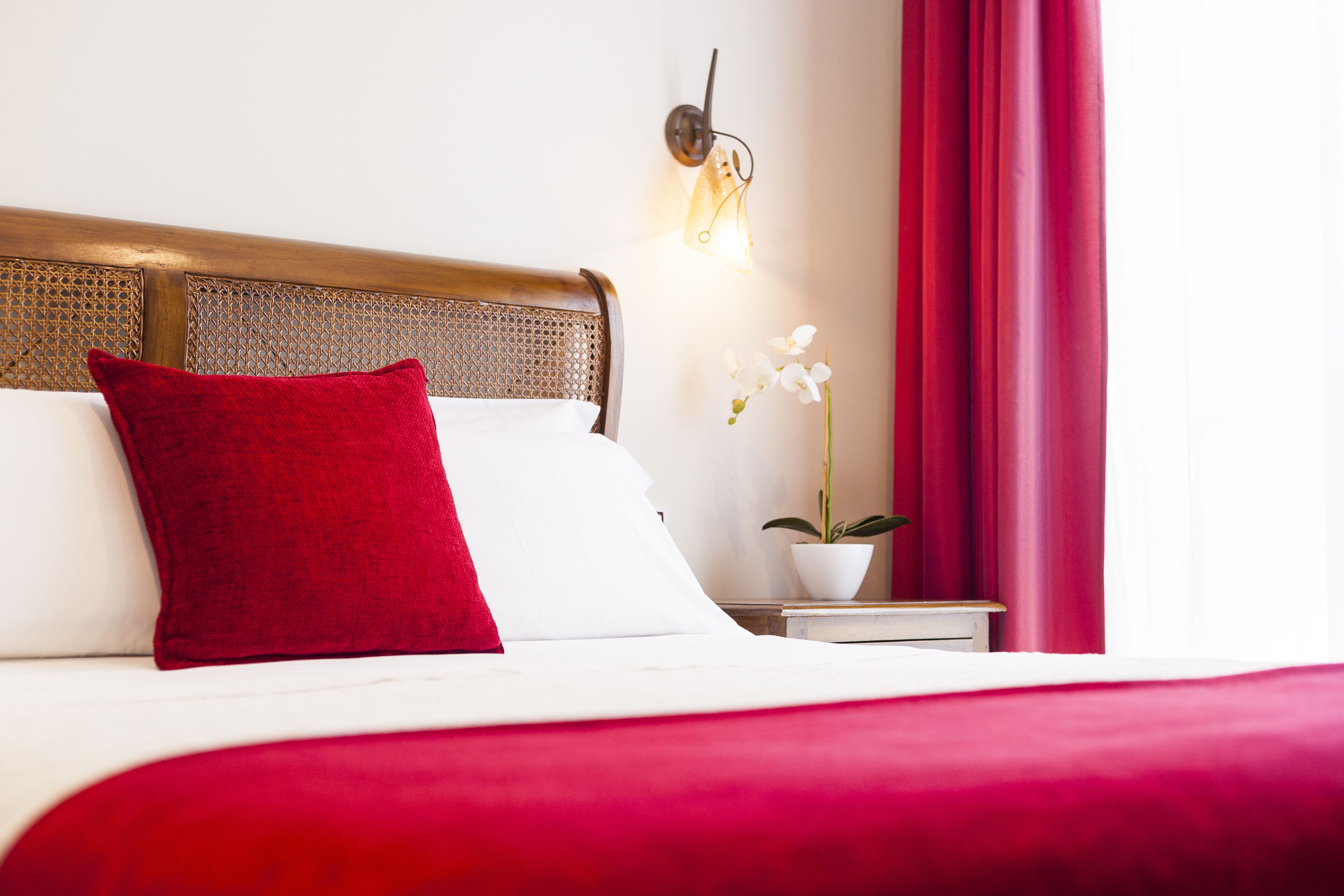 A neatly made bed with a red pillow, a white pillow, and a red bedspread. There is a small bedside table with a white lamp and a potted plant.