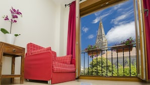 A red sofa, a wooden side table, and a potted orchid in a room with a view of a building through a glass door.