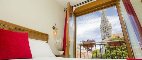 A hotel room with a large window, a bed with white linens, a red pillow, and a view of a tower.