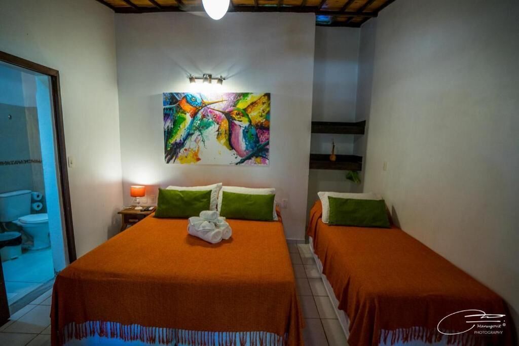 Habitación de hotel con dos camas, un cuadro abstracto de colores sobre ellas, una mesita de noche con una lámpara, y un baño visible a la izquierda.