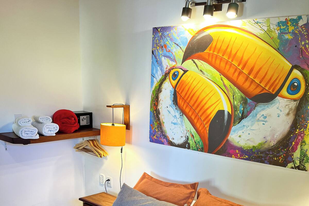 A neatly made bed with a brown blanket, a headboard with pillows, a wall-mounted painting of a toucan, a shelf with towels and a lamp, and a bedside table with a book and a phone charger.